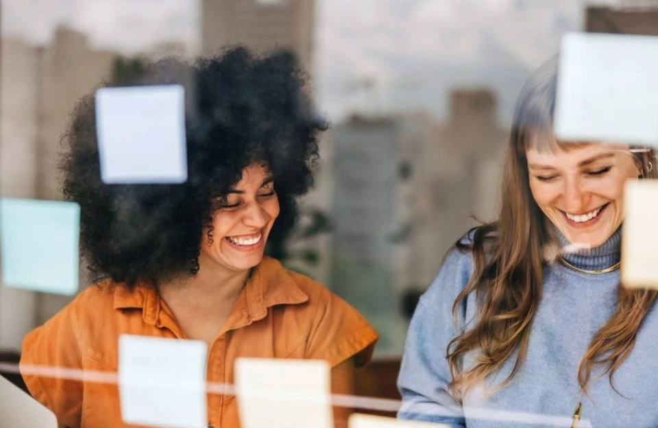 Two women smiling and collaborating in an office, viewed through a glass wall with colorful sticky notes. The atmosphere is cheerful and creative.