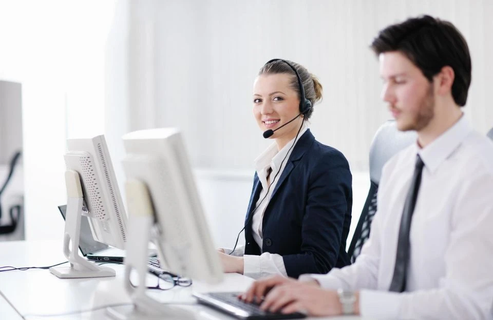 Two call center workers are sitting at desks. The woman on the left, wearing a headset and suit, is smiling at the camera. Both work on computers.