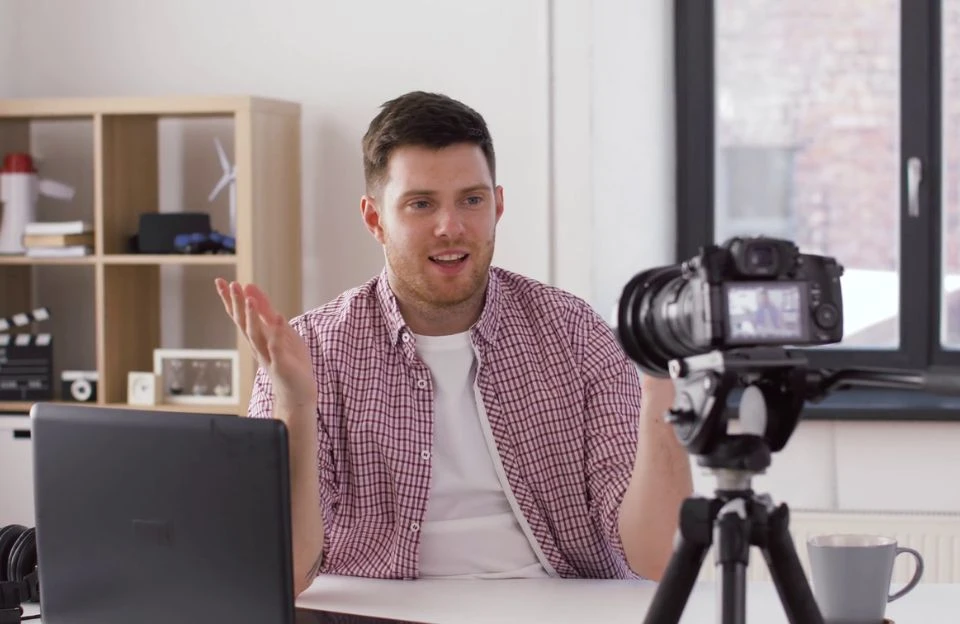 A man in a casual checkered shirt gestures while speaking in front of a camera on a tripod. A laptop and headphones are on the table. The room is bright, with shelves and a window in the background